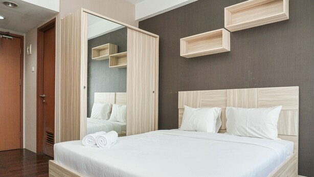 A neatly made bed with white linens and pillows, a wooden headboard, and a large mirror with a wooden frame. - Simply Studio Room Apartment Margonda Residences 5 (Depok)