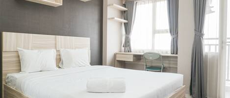 A neatly made bed with white linens and a folded towel, a desk with a chair, and a window with curtains.