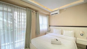 A hotel room with a large bed, a nightstand, an air conditioning unit, and sheer curtains. - Cozy and Stylish 1BR Apartment at Gateway Pasteur (Cimahi)