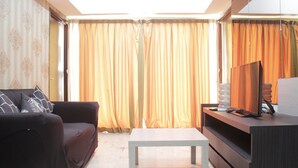A living room with a sofa, a coffee table, a TV on a wooden stand, and a large window with curtains. - Deluxe & Comfy 2BR at Braga City Walk Apartment (Bandung)