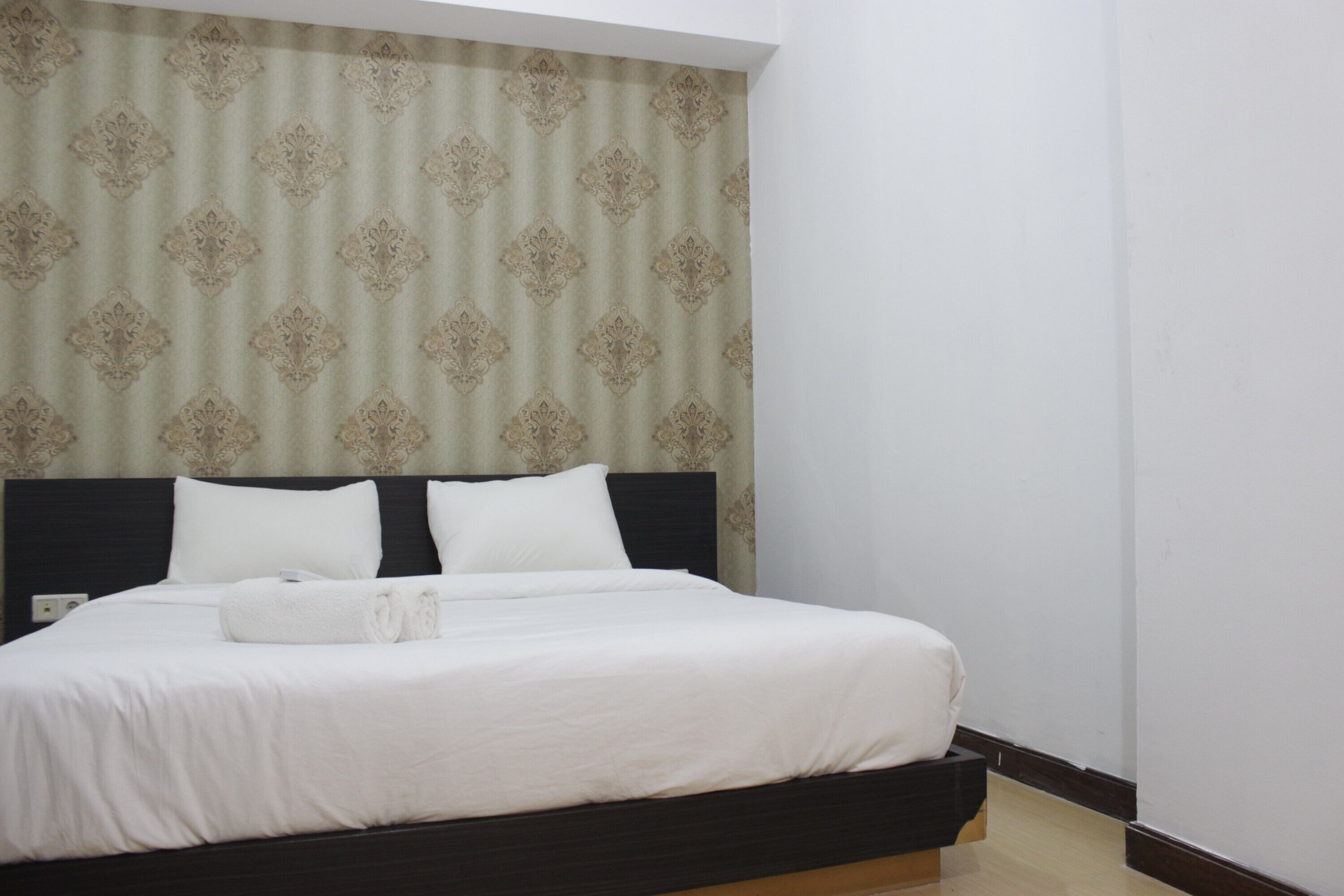 A neatly made bed with white linens and two pillows, a dark headboard, and a patterned curtain in the background.