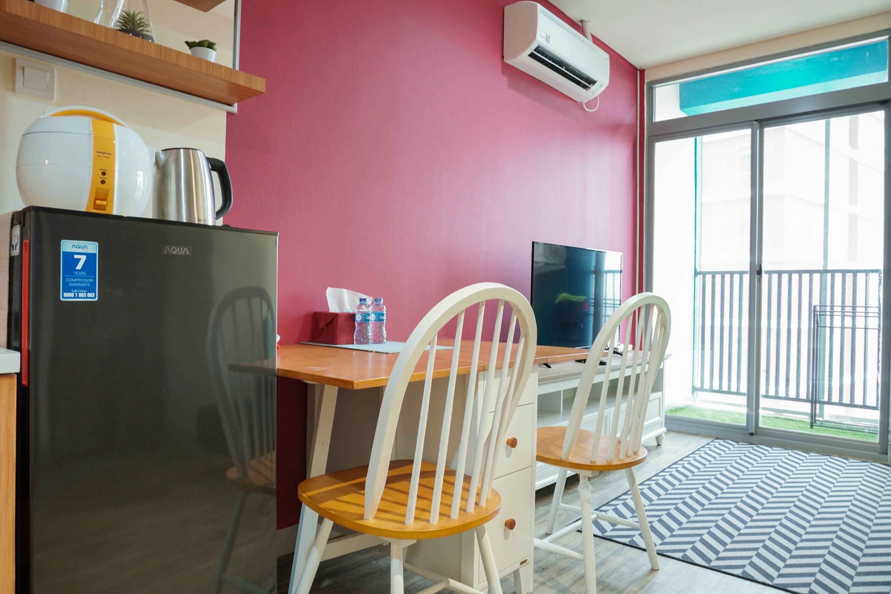A compact kitchenette with a refrigerator, a kettle, and a coffee maker. There are two white chairs and a wooden table. The room has a red wall and a large window with a view of a balcony.