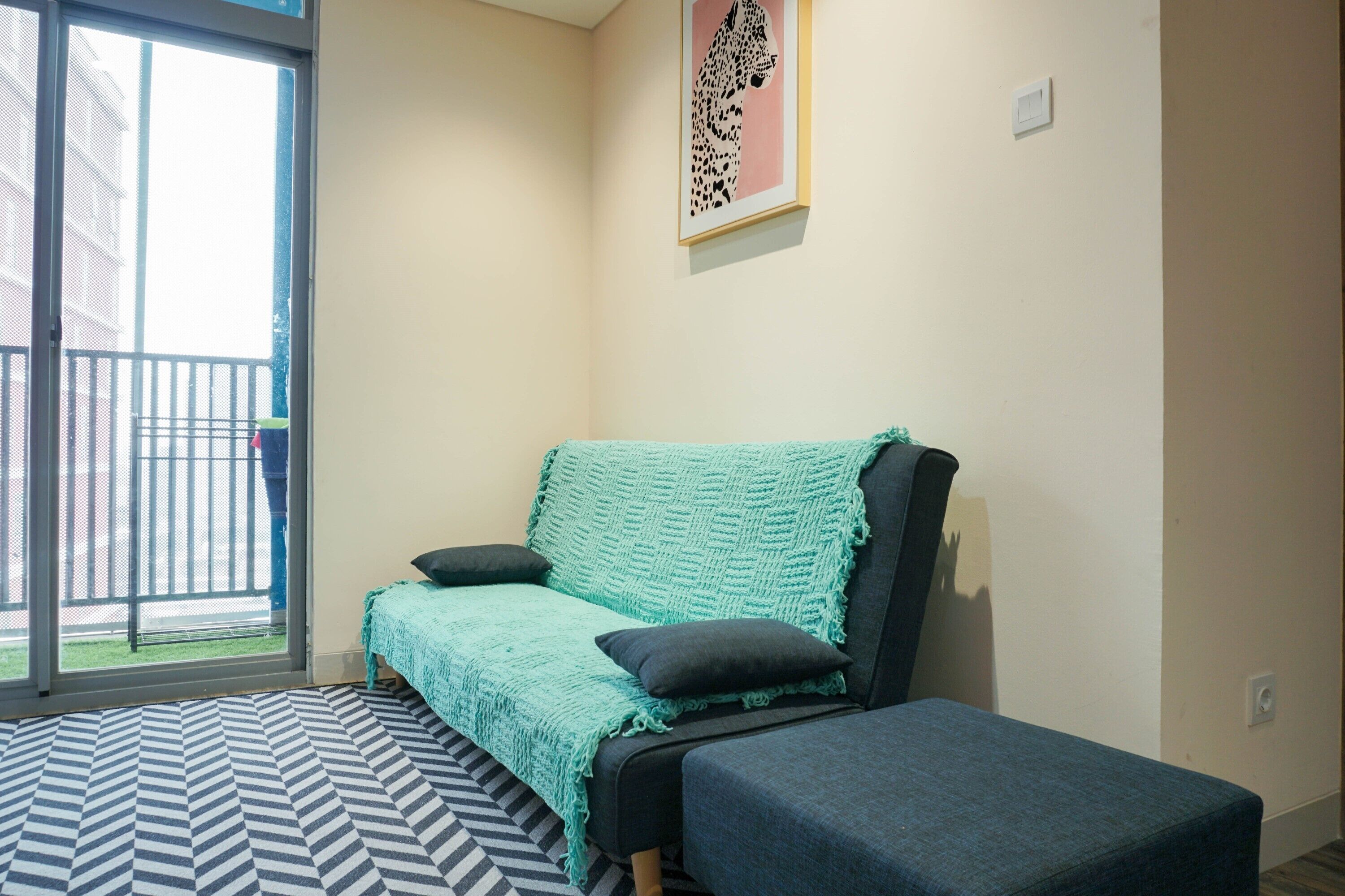 Una habitació amb un sofà i un reposapius, una catifa estampada, una obra d'art emmarcada a la paret i una porta corredissa de vidre que dona a un balcó.