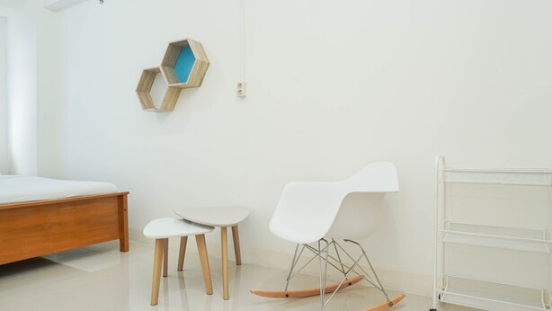 A modern room with a wooden bed, two white chairs, a rocking chair, and a white metal shelf unit. - Cozy and Simply 1BR Green Pramuka Apartment (Jakarta)