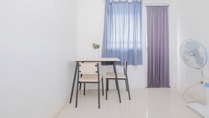 A small room with a table and two chairs, a window with blue and purple curtains, and a standing fan. - Cozy and Simply 1BR Green Pramuka Apartment (Jakarta)