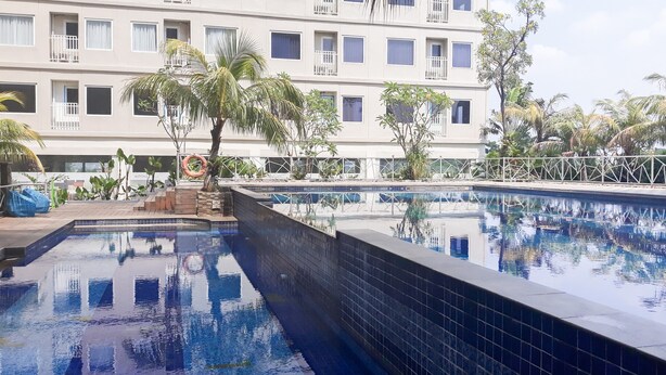 Une piscine dont le bord est recouvert de carreaux, entourée de palmiers et bordée d’un immeuble doté de nombreux balcons.