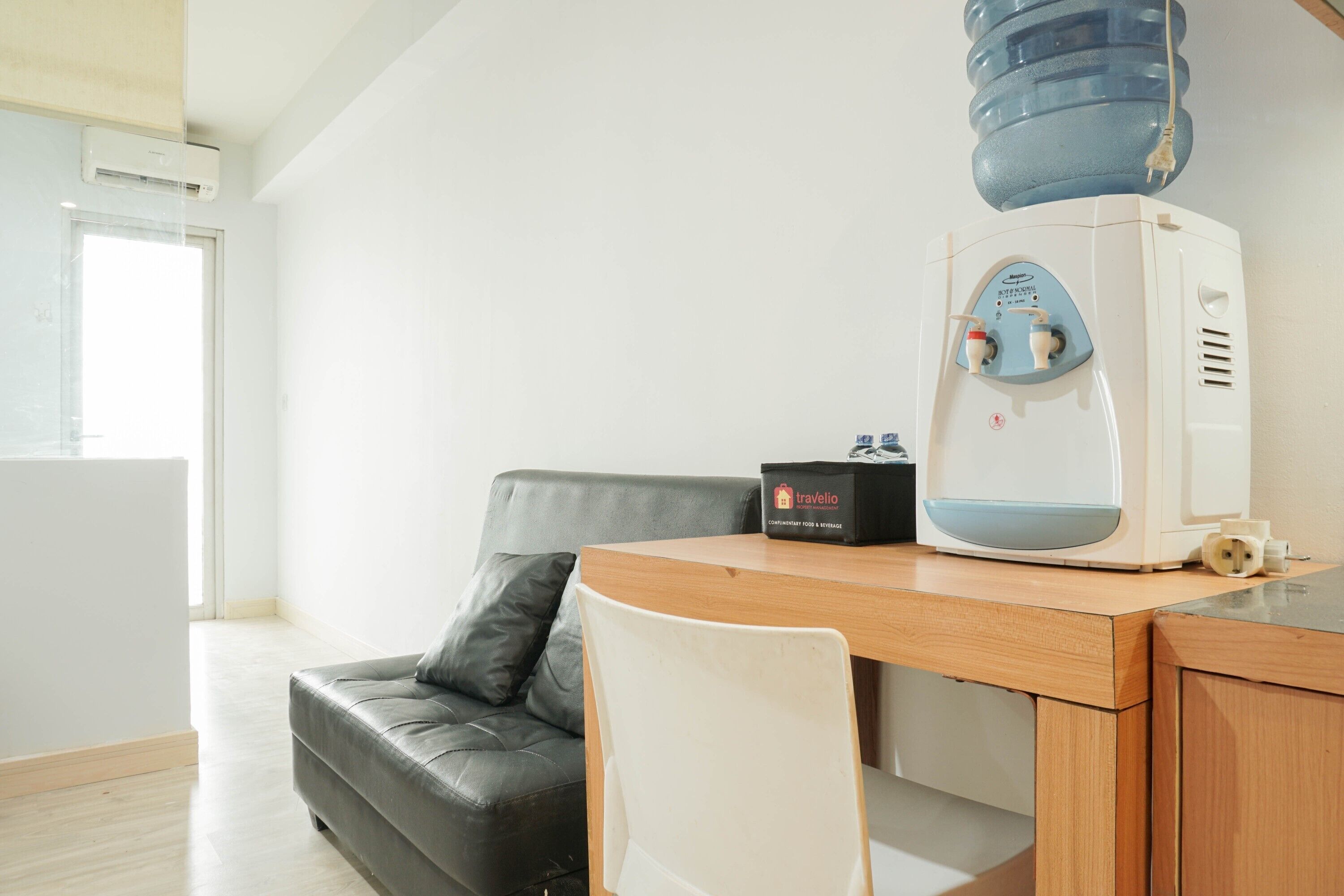 A modern living room with a grey leather sofa, a wooden table, a water dispenser, and a travel bag on the table.