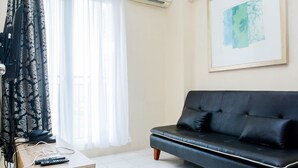 A living room with a black leather sofa, a wooden coffee table with remote controls, and a window with sheer curtains. - Comfy 2BR Mutiara Bekasi Apartement (Bekasi)