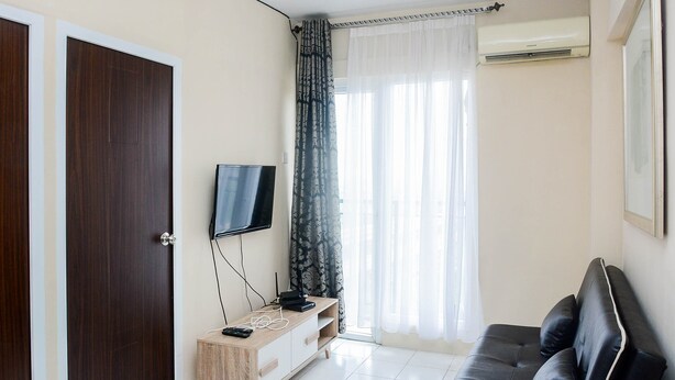 A living room with a dark sofa, a wooden TV stand, a flat-screen TV, and a window with sheer curtains. - Comfy 2BR Mutiara Bekasi Apartement (Bekasi)