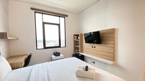 A hotel room with a large bed, a TV mounted on a wooden panel, a small desk with a chair, and a window with a city view.
