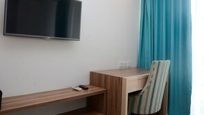 A hotel room with a flat-screen TV mounted on the wall, a wooden desk with a chair, and a window with teal curtains.