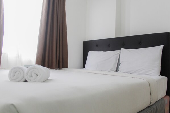 A neatly made bed with white linens and two rolled towels, a dark headboard, and brown curtains.