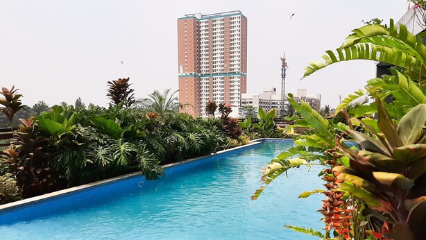 A swimming pool with clear blue water, surrounded by lush tropical plants and a tall residential building in the background. - Minimalist and Cozy Studio Apartment at Tuscany Residences (South Tangerang)