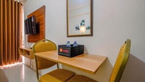 A hotel room with a wooden desk, two yellow patterned chairs, a flat-screen TV, a mirror, and a minibar with a travel logo. - Comfy Studio at Bogorienze Apartment near The Jungle Waterpark (Bogor)
