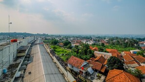 A high vantage point overlooking a town with red-roofed houses, a clear sky, and a distant view of a larger city.