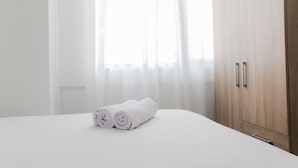 Ein ordentlich bezogenes Bett, gerollte weiße Handtücher, ein Holzschrank und durchscheinende Vorhänge, die natürliches Licht hereinlassen.