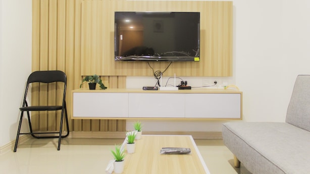 Ein modernes Wohnzimmer mit einem an der Wand montierten Flachbildfernseher, einem schwarzen Klappstuhl, einem grauen Sofa, einer weißen Kommode und einem kleinen Couchtisch mit Topfpflanzen.
