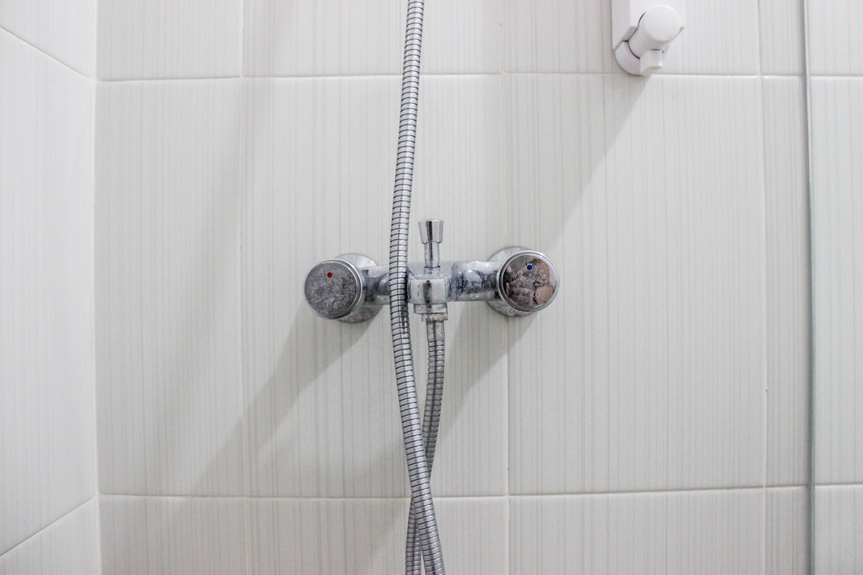 A shower with a hose and two faucets against a tiled wall.
