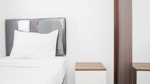 Una cama bien hecha con cabecera gris y ropa de cama blanca, una mesita de noche de madera a un lado y una puerta de madera a la derecha.
