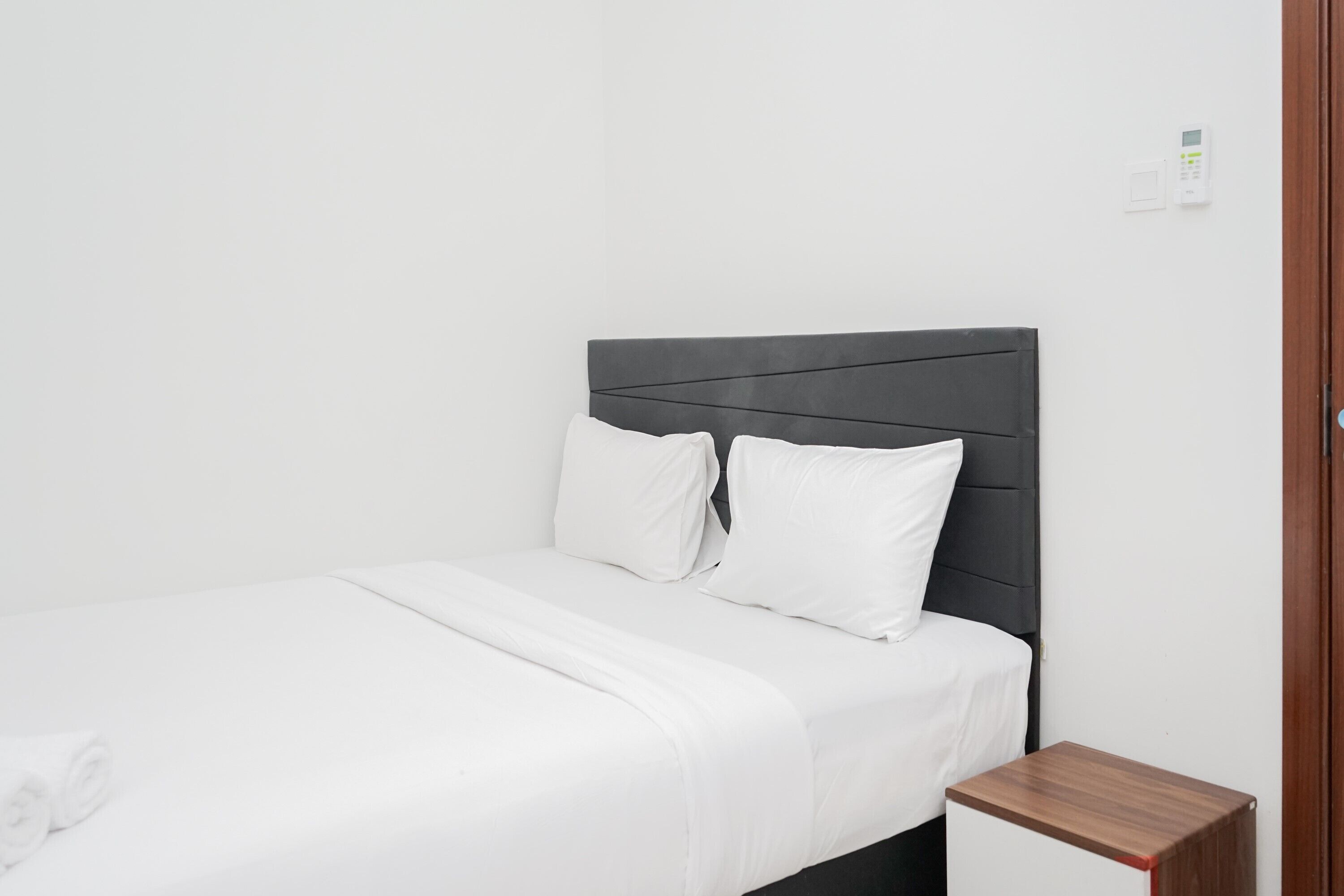 Un lit bien fait, avec du linge de lit blanc et des oreillers, une petite table de chevet en bois à côté, et un mur blanc sobre agrémenté d’un thermostat.