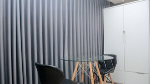 Une pièce avec une table dont le plateau est en verre et deux chaises noires, une armoire blanche et des rideaux gris.