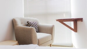 A beige armchair with a zigzag pillow, a small wooden side table, and a wooden shelf against a white wall with blinds partially open.