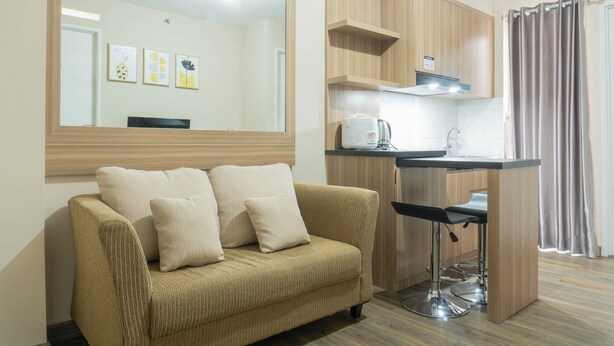 A modern living room with a beige sofa, a coffee table, and a large mirror. There is a kitchenette with a stove, sink, and bar stools.