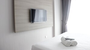 Ein modernes Hotelzimmer mit einem an einer Holzvertäfelung montierten Flachbildfernseher, einem ordentlich bezogenen Bett und weißen Handtüchern, sowie einem Fenster mit grauen Vorhängen.