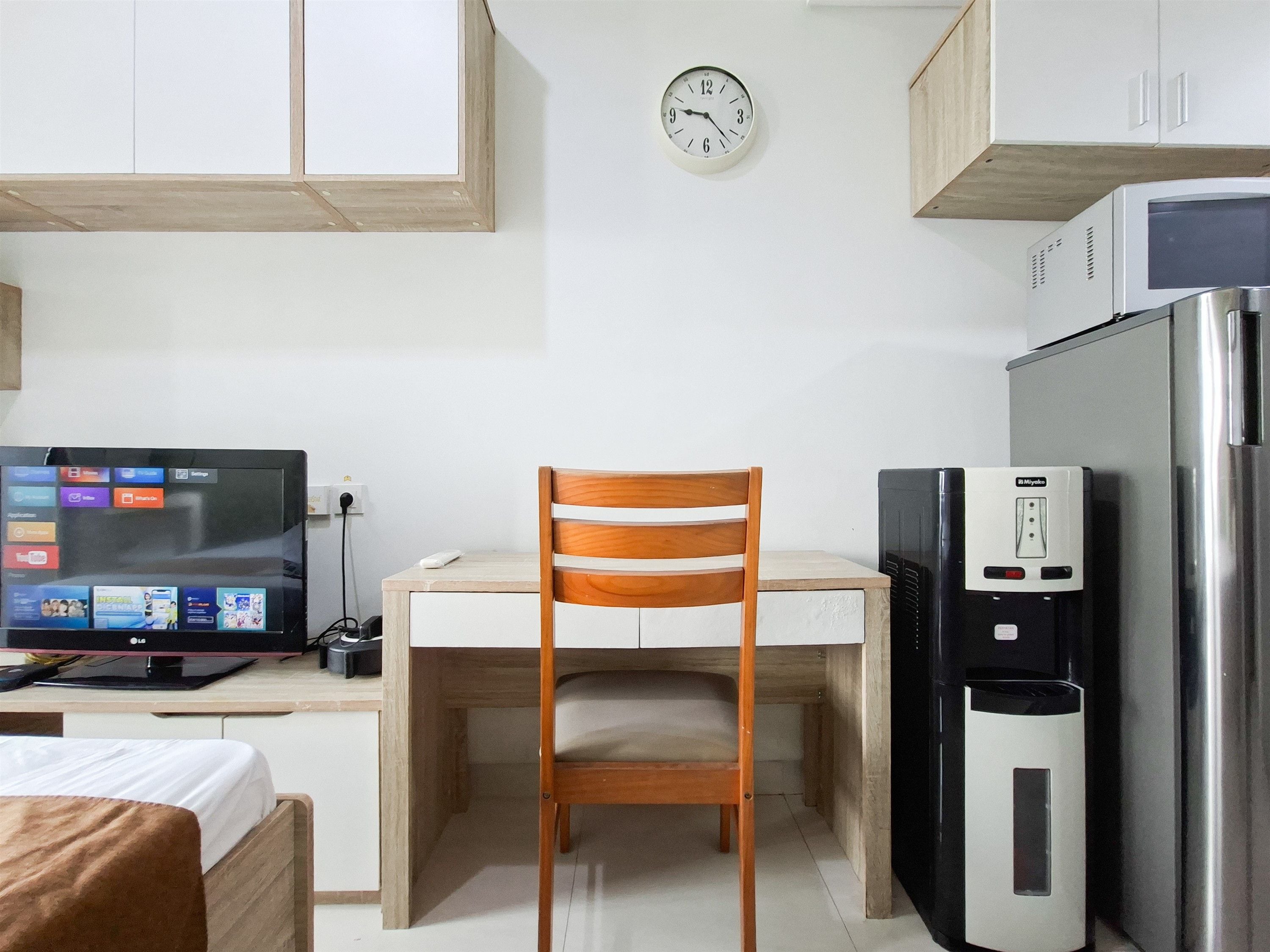 A compact kitchenette with a wooden chair, a refrigerator, a microwave, and a TV displaying a home screen.