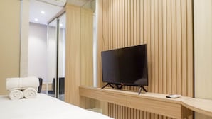 A modern hotel room with a large bed, a flat-screen TV mounted on a wooden panel wall, and a desk with a remote control. - Best Studio Benson Apartment Connected to Pakuwon Mall next to Anderson (Surabaya)