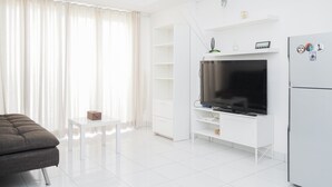 A modern living room with a flat-screen TV, white furniture, and a large window with curtains.