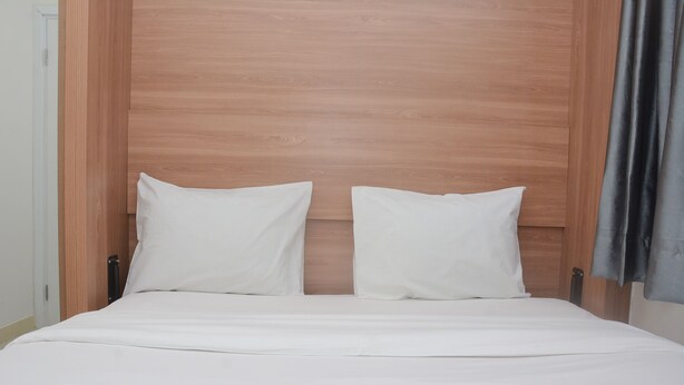 A neatly made bed with white bedding and pillows, a wooden headboard, and a grey curtain on the right.