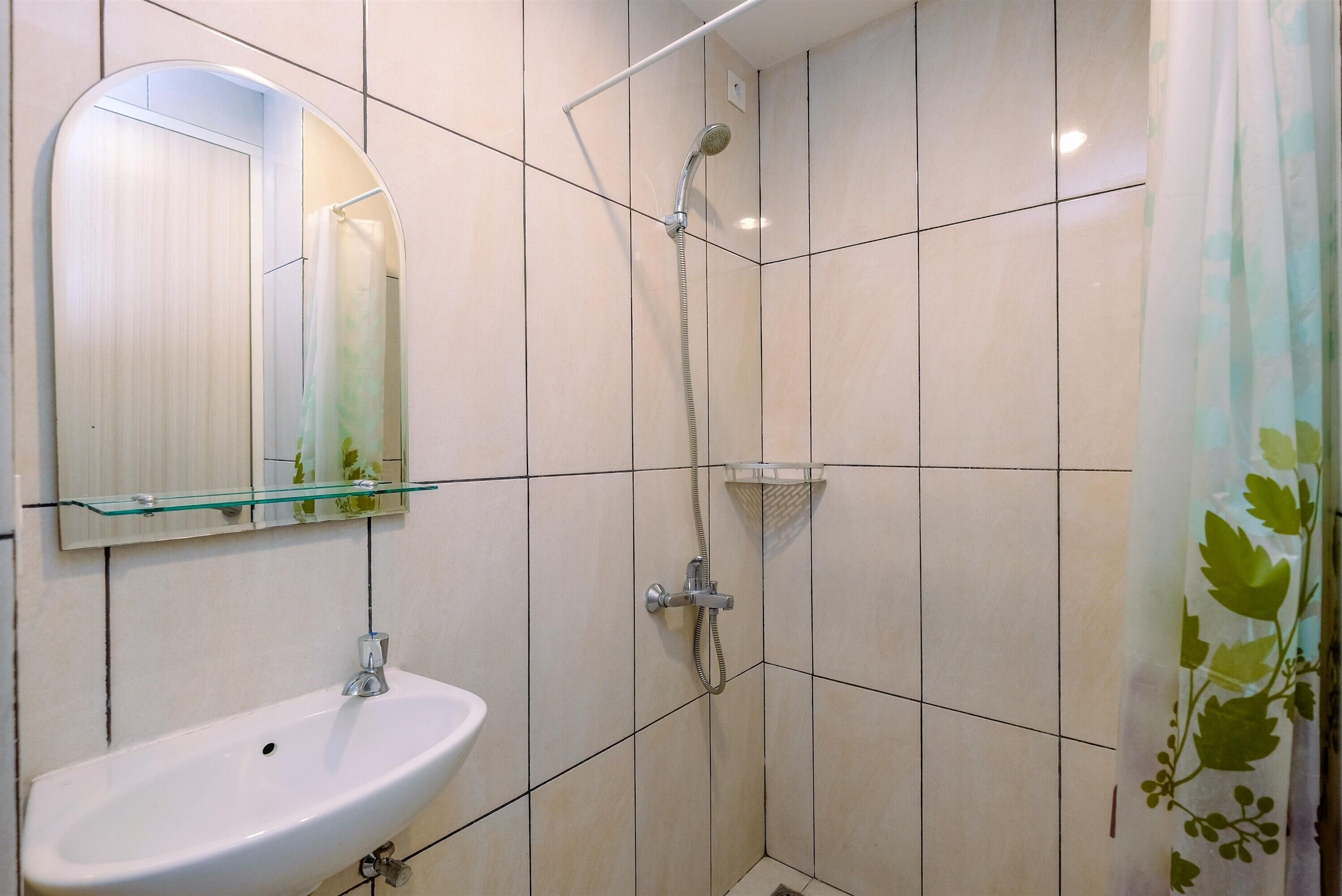 A bathroom with a white sink, a glass shelf above it, a shower with a handheld showerhead, and a mirror with a leaf pattern.