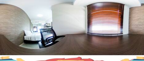 A modern hotel room with a curved wooden floor, a bed with white linens, a black chair, and a large window with blinds.