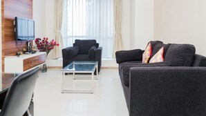 Ein modernes Wohnzimmer mit einem dunkelgrauen Sofa, einem Glastisch, einer Holz-TV-Konsole und einem Fenster mit transparenten Vorhängen.