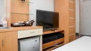 Une chambre à coucher de taille modeste, comprenant un lit, un téléviseur, un petit réfrigérateur, une armoire en bois et une étagère avec une plante.