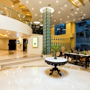A spacious lobby with a marble floor, a central column with a green leafy pattern, and a seating area with tables and chairs.