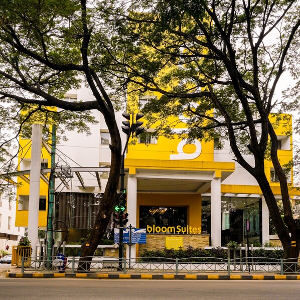 A yellow building with 'bloom suites' signage, surrounded by trees and a gated entrance.