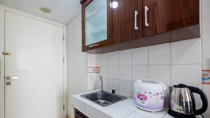 A small kitchen with white tiles, a stainless steel sink, wooden cabinets, a white and purple rice cooker, and a black electric kettle. - Comfortable Studio at Springlake Summarecon Bekasi Apartment (Bekasi)