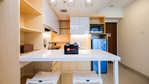 A modern kitchen with white cabinetry, a white countertop, and a travel bag on the counter. There are two white chairs under the counter and a microwave above the counter. - Spacious Studio at Grand Kamala Lagoon Apartment (Bekasi)