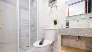 A modern bathroom with a glass-enclosed shower, a white toilet with a towel on it, a sink with a glass shelf, and a marble wall. - Spacious Studio at Grand Kamala Lagoon Apartment (Bekasi)