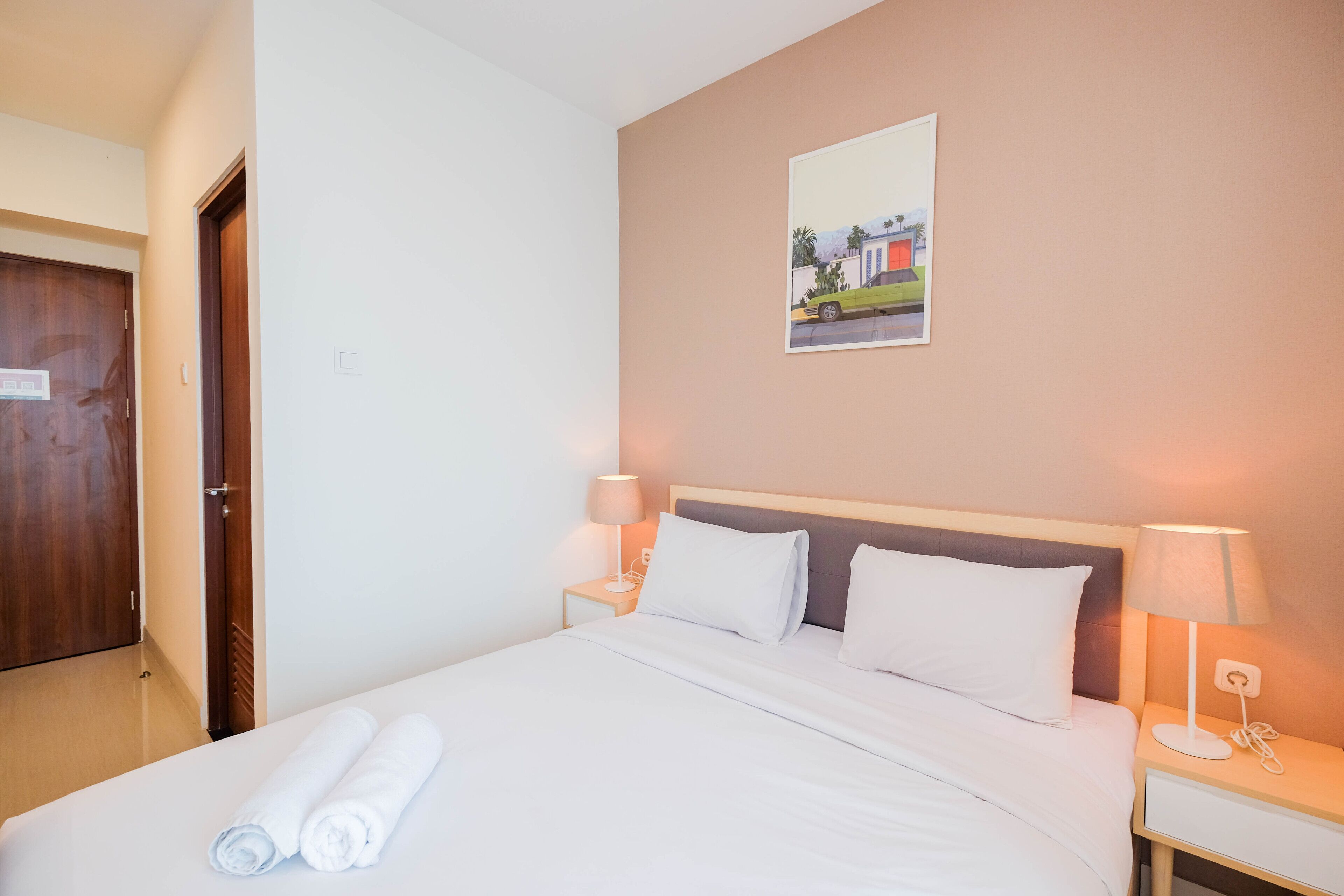 A neatly made bed with white linens and rolled towels, a wooden door, a nightstand with a lamp, and a wall-mounted picture.