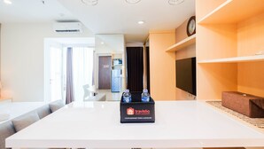 A hotel room with a white table, a sofa, a TV on the wall, and a clock. There is a minibar with bottles and a tray. - Spacious Studio at Grand Kamala Lagoon Apartment (Bekasi)