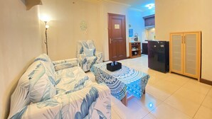 Un salon comprenant un canapé, un pouf à motifs, un fauteuil et une porte donnant sur un espace cuisine.