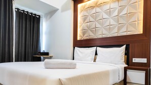 A neatly made bed with white linens and a rolled towel, a wooden headboard with geometric patterns, a bedside table with a lamp, and a power outlet on the nightstand.