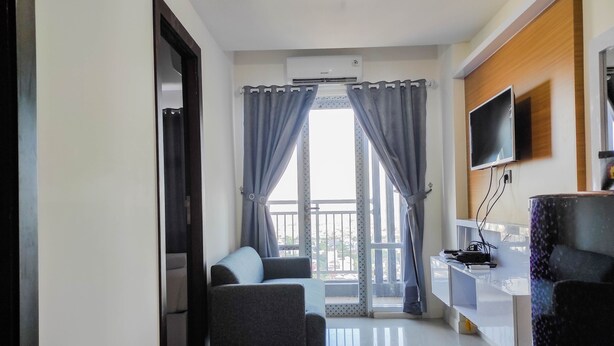 A modern living room with a sofa, a TV mounted on the wall, and a balcony with curtains. - Homey and Comfy 2BR at Vida View Apartment (Makassar)
