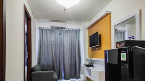 A modern hotel room with a grey sofa, a flat-screen TV mounted on a wooden panel, a black refrigerator, and a bathroom with a shower curtain. - Homey and Comfy 2BR at Vida View Apartment (Makassar)