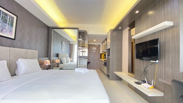 A modern hotel room with a large bed, a TV mounted on the wall, and a kitchen area visible through an open door. - Luxurious Studio Room at Galeri Ciumbuleuit 3 near UNPAR (Bandung)