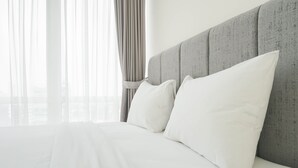 Cama bem arrumada com lençóis e almofadas brancas, cabeceira estofada cinza e cortinas leves que permitem a entrada de luz natural.
