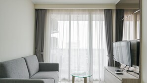 Uma sala de estar moderna com um sofá cinzento, uma mesa de centro de vidro, uma televisão de tela plana e cortinas leves.